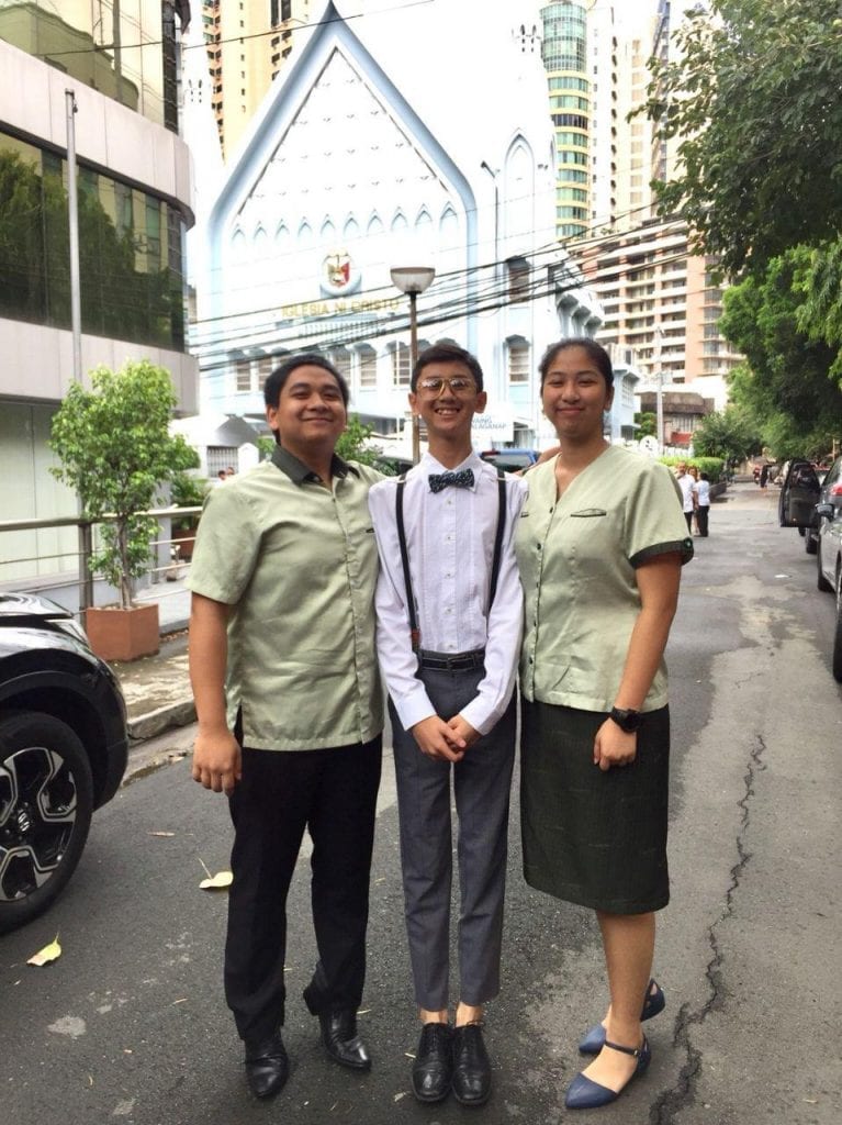 A group of 3 young people standing on the street with an INC house of worship behind them.