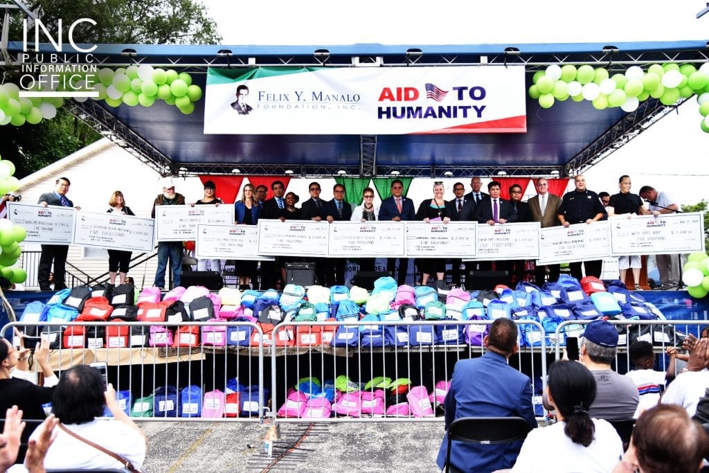 Crowd on a stage holding 10 large donation checks; hundreds of backpacks of school supplies in front of stage