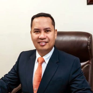 minister wearing suit and orange tie, smiling and sitting in brown chair